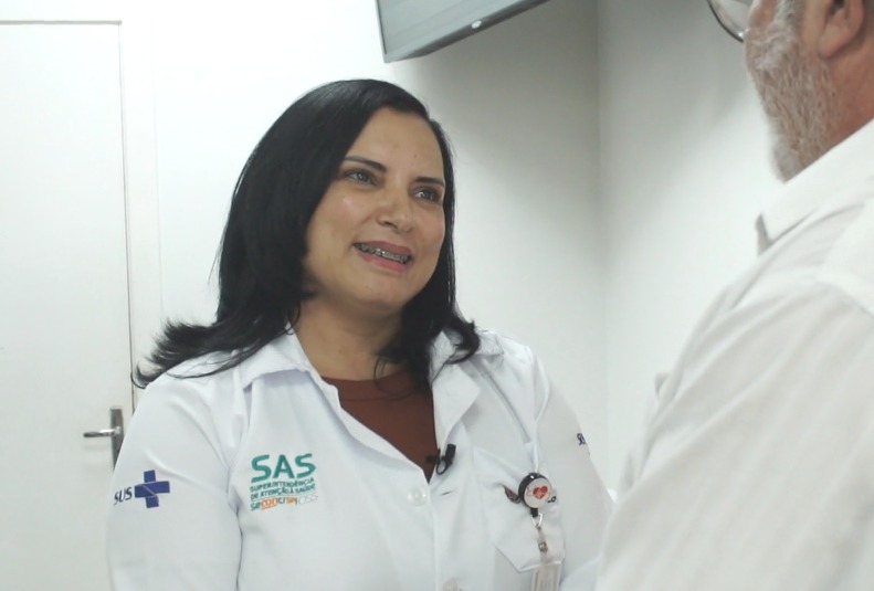Na imagem, Marcia, uma mulher de 43 anos, cabelo preto e comprido e aparelho nos dentes, usa jaleco branco com logotipo do SUS e está sorrindo para uma pessoa à sua frente, um homem que aparece de perfil e usa camisa branca, óculos e tem barba branca. Os dois estão em uma sala de paredes brancas